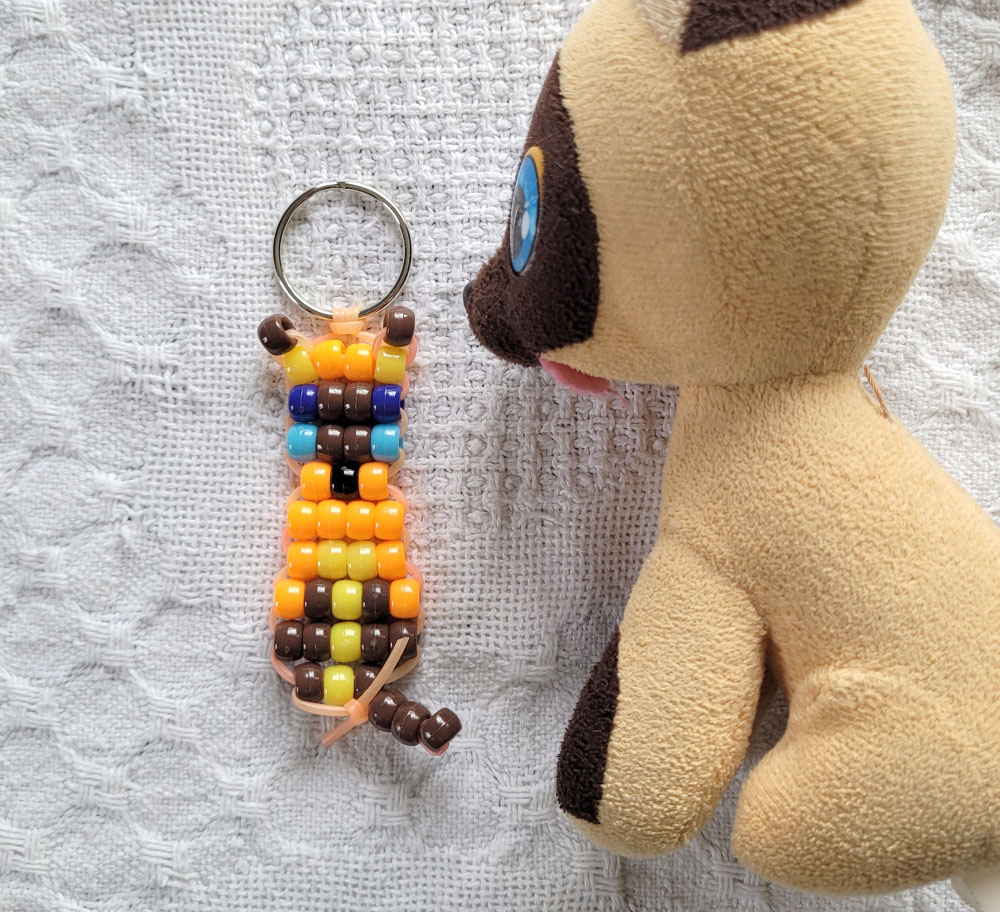 A pony-bead keychain of an orange siamese kitten, next to a plush toy of the same cat