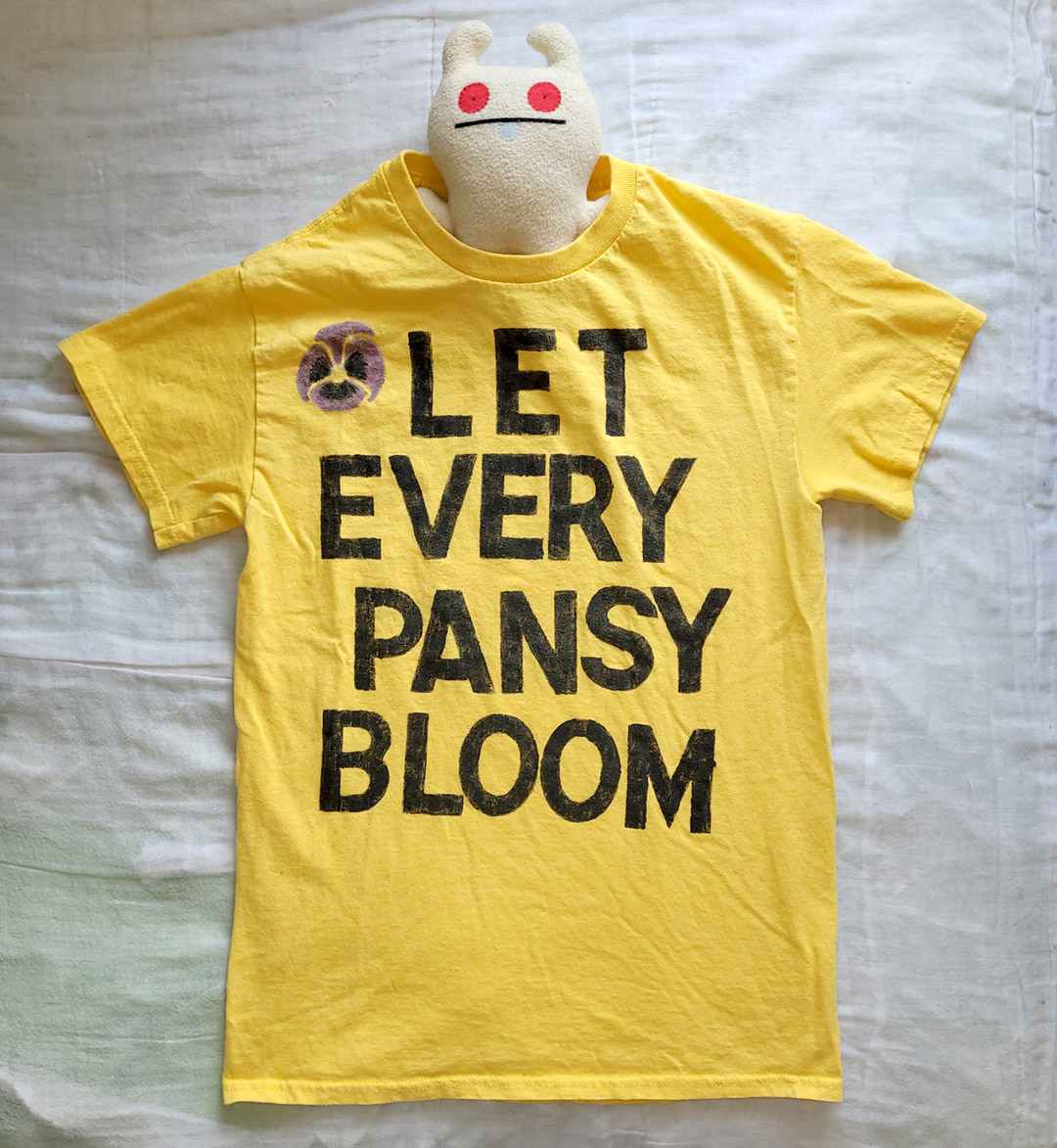 A yellow shirt with black text and a small purple pansy, reading Let Every Pansy Bloom