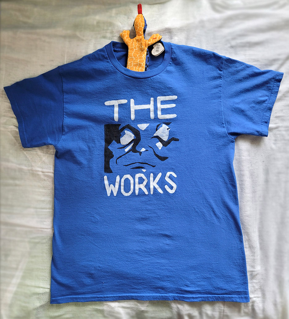 A blue shirt with a black and white face on it, reading The Works