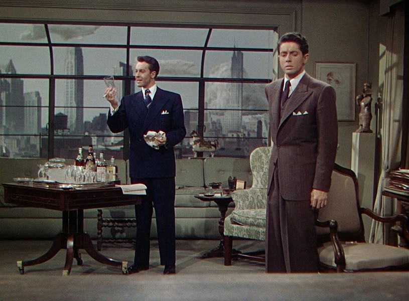 Two men standing in a living room; one cleaning a glass, the other staring in an uncomfortable horror at something.